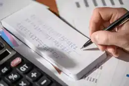 Mão a anotar valores financeiros num bloco junto a uma calculadora
